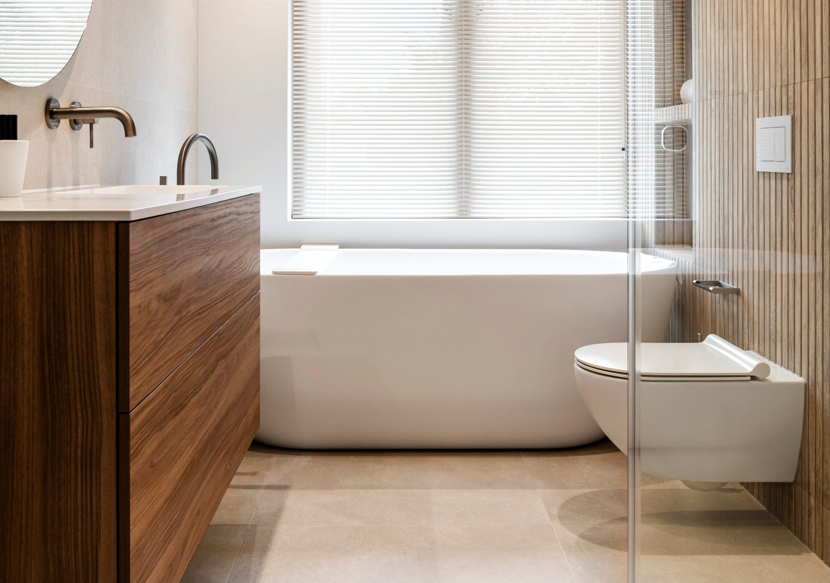 Scandinavische badkamer met een strakke houten wastafelkast, een witte marmeren bovenkant, wandgemonteerde kraan, ronde spiegel boven de wastafel en een witte vrijstaande badkuip bij het raam met jaloezieën.